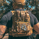 Back view of Male wearing a tactical plate carrier with Slav Squat morale patch velcroed to front chest while holding an AR-style rifle. Also wearing TAPR with P-CAN