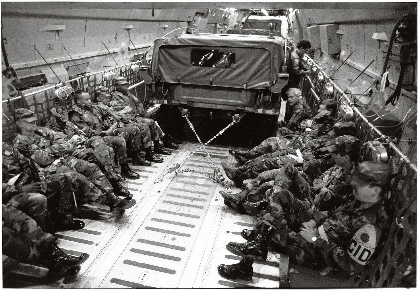 Black-and-white photo of U.S. Army soldiers seated inside a military cargo aircraft with a Humvee secured by chains during troop transport.
