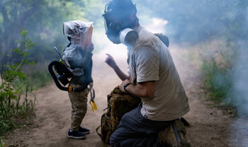 A man wearing MIRA Safety CM-6M Gas mask kneels on a smoky path, guiding a young child wearing MD-2™ CBRN Kids Gas Mask Respirator with PAPR.