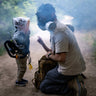 A man wearing MIRA Safety CM-6M Gas mask kneels on a smoky path, guiding a young child wearing MD-2™ CBRN Kids Gas Mask Respirator with PAPR.