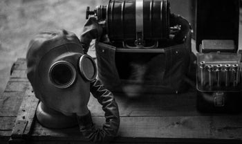 Vintage gas mask with breathing hose, filtration device, and ammunition box on a wooden crate in black and white.