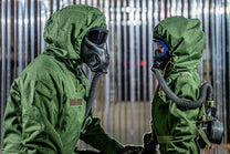 Two individuals in green protective suits with gas masks against a metallic background