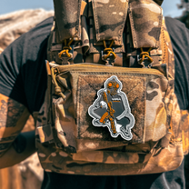 Close up Back view of Male wearing a tactical plate carrier with Slav Squat morale patch velcroed to front chest while holding an AR-style rifle. Also wearing TAPR with P-CAN