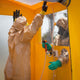 A man in a hazmat suit being sprayed with a cleansing solution. A man from the outside of the shower is using a spray gun through a glove system.