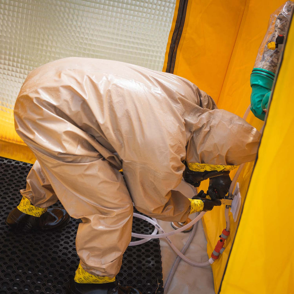 A man wearing a hazmat suit inside of the decontamination shower is tightening the valve switches. The drainage mat can also be seen.